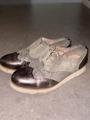 Gråa sneakers från Tom Tailor - Snygga sneakers från Tom Tailor i grå mocka och metallicdetaljer. Skorna har klassisk snörning, perforerade mönster och vit sula. Perfekt för dig som gillar en mix av stilrent och trendigt.