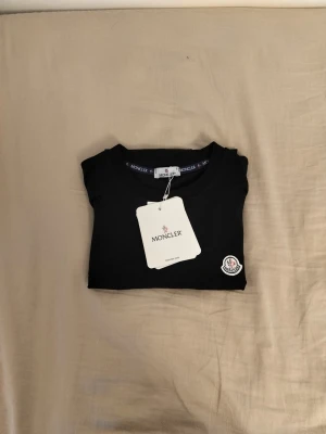 Svart Moncler t-shirt i bomull - Stilren svart t-shirt från Moncler med klassisk rund hals och korta ärmar. T-shirten har Monclers ikoniska logga broderad på bröstet och är tillverkad i mjuk bomull för en skön känsla. Perfekt till jeans eller shorts.