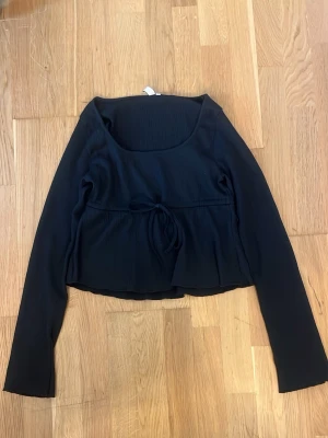 Svart långärmad topp med knyt - Trendig svart långärmad topp med rundad urringning och knyt i midjan. Toppen har en croppad passform och är tillverkad i ett mjukt, stretchigt material som ger en bekväm känsla. Perfekt att styla med jeans eller kjol.
