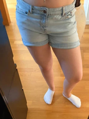 Ljusblå denimshorts med uppvik - Säljer ett par ljusblå denimshorts med klassisk femficksmodell och uppvikta benslut. Shortsen har normal passform och midja, samt bälteshällor och knappgylf. Perfekta för varma dagar och enkel att matcha med olika toppar. Skriv pm vid funderingar!☺️