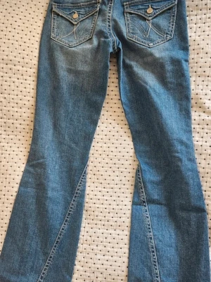Blå slitna bootcut jeans - Säljer ett par ljusblå bootcut jeans med slitningar och hål på framsidan av benen. Jeansen har två bakfickor med dekorativa sömmar och knappdetaljer. Märket är Gina Tricot, storlek 164