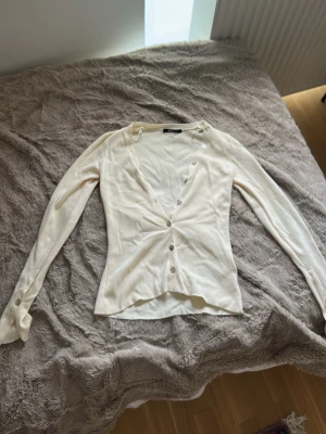 Vit ribbad kofta med knappar gina tricot - Säljer en stilren vit kofta med ribbad struktur och v-ringning. Koftan har långa ärmar och knäpps med silverfärgade knappar framtill. Materialet känns mjukt och stretchigt, perfekt för lager på lager. Enkel och snygg att matcha till olika outfits. Aldrig använd!!