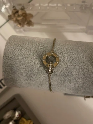 Edblad armband  - Snyggt armband i guldton med en rund cirkel i mitten, dekorerad med små gnistrande stenar. Kedjan är tunn och elegant, och cirkeln har små detaljer runt om. Perfekt för dig som gillar stilrena accessoarer med lite bling.