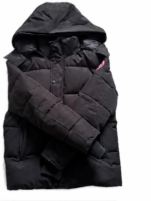 Svart dunjacka från Canada Goose - Säljer en svart dunjacka från Canada Goose med huva och klassisk patch på ärmen. Jackan har en puffig design, dragkedja och tryckknappar framtill samt ribbade muddar vid ärmsluten. Perfekt för kalla vinterdagar och riktigt varm. Det står Xs men de kommer passa dig som har storlek M
