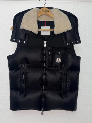 Svart dunväst från Moncler - Snygg svart dunväst från Moncler med avtagbar huva och krage i beige fuskpäls. Västen har dragkedja framtill, två stora fickor med dragkedja och en bröstficka med knapp. Klassisk Moncler-logga på bröstet och quiltad design för extra värme.
