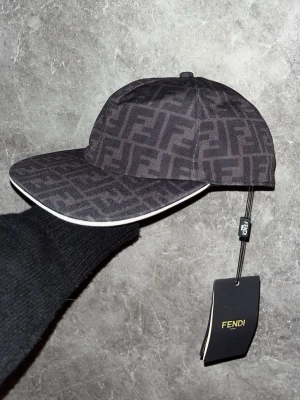 FENDI KEPS - Snygg Fendi-keps med det klassiska FF-mönstret i svart/brunt. Stilren modell med vit detalj längs skärmen. Kommer med originaltags kvar, vilket gör den extra fin för försäljning.  * Fendi FF-mönster * Svart/brun * Vit detalj på skärmen * Originaltags kvar * Mycket fint skick