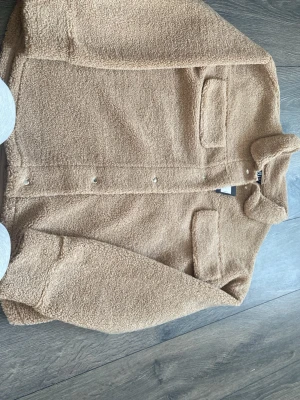 Beige teddyjacka med fickor - Mysig beige teddyjacka i fluffigt material med tryckknappar framtill och två bröstfickor med lock. Jackan har en avslappnad passform och mjuk struktur som ger en trendig look. Perfekt för dig som gillar streetstyle och vill hålla dig varm.