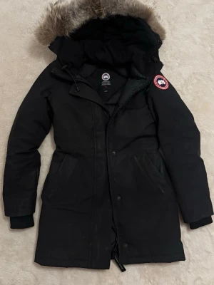 Svart lång dunjacka med pälshuva - Säljer en svart lång dunjacka från Canada Goose med avtagbar huva och snygg pälskant. Jackan har två stora fickor framtill, dragkedja och knappar samt den klassiska loggan på ärmen. PASSAR XS då den inte är liten i strlkn PRIS KAN DISKUTERAS 