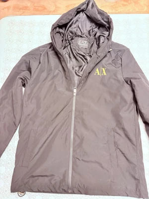  vindjacka (Emporio Armani,A|X) - Snygg brun vindjacka från Emporio Armani EA7 med huva och dragkedja framtill. Jackan har två sidofickor och gul A|X-logga på bröstet. Tillverkad i lätt och glansigt material som passar perfekt för blåsiga dagar.