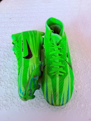 Nike gröna Mercurial fotbollsskor - Säljer ett par grymma Nike Mercurial fotbollsskor i neon-grönt med svarta detaljer och blå inslag. Skorna har snörning, mönstrad ovandel och dobbar för bra grepp på planen. Perfekta för dig som vill sticka ut på fotbollsplanen!