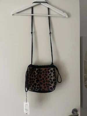 Leopardmönstrad pärlhandväska - Snygg handväska med leopardmönster i pärlor i svart, beige och brunt. Väskan har en justerbar axelrem och dragskostängning. Sidorna är i svart tyg som ger en cool kontrast till det glittriga mönstret. Perfekt för dig som vill sticka ut med din stil.