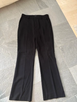 Svarta kostymbyxor med raka ben - Snygga svarta kostymbyxor med hög midja och raka ben. Byxorna har bälteshällor och sidofickor, vilket ger en clean och stilren look. Perfekta för dig som gillar klassisk stil med modern touch.