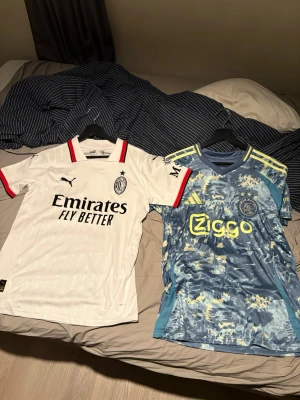 Två fotbollströjor Puma och Adidas - Säljer två snygga fotbollströjor: en vit AC Milan-tröja från Puma med svarta och röda detaljer samt en blå Ajax-tröja från Adidas med gula och ljusblå mönster och gula detaljer. Perfekta för match eller träning!
