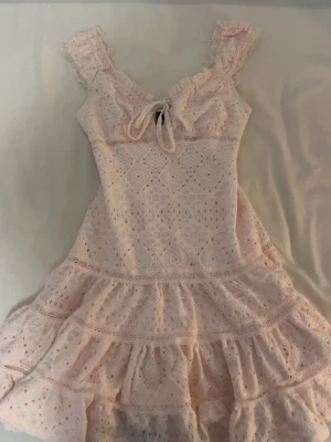 Ljusrosa spetsklänning Gina Tricot - Supersöt ljusrosa kort klänning från Gina Tricot med volanger, spetsdetaljer och knyt framtill. Klänningen har breda axelband med volang och ett romantiskt mönster. Perfekt för sommarens alla tillfällen!