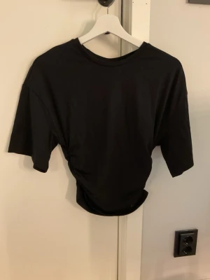 Svart topp från ginatricot - En svart topp med korta ärmar och cool snörningsdetalj i sidan som ger en snygg twist. Tillverkad i mjuk bomull och har en relaxed fit. Perfekt för dig som gillar enkla men ändå unika plagg.