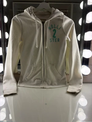 Hollister Vintage Zip Up Hoodie i Off-white / Cream (Y2K) i strl xs - Säljer denna jättesnygga zip-up hoodie från Hollister i en fräsch off-white/krämvit färg! 😍Tröjan har ett superfint turkost tryck framtill med texten "HOLLISTER" och en stor tvåa som är detaljerad med lite diskret, fint glitter ✨ Perfekt för en mysig men trendig Y2K-look.               Märke: Hollister Co.                                             Färg: Off-white / Cream med turkosa detaljer.   Skick: Mycket bra skick / Sparsamt använd.              Storlek: xs