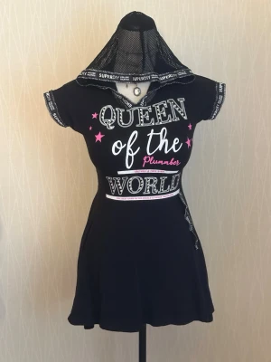 Svart kortklänning med text och meshhuva - Svart kortklänning med vit och rosa text 'Queen of the World' och stjärndetaljer. Klänningen har meshhuva, korta ärmar och dekorativa band med texten 'Superdiy College Athletics' längs huva och midja. Materialet ser ut att vara polyester eller syntet.