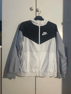 Nike vit och svart vindjacka med huva - Nike vindjacka i vitt och svart med dragkedja framtill och huva. Jackan har två sidofickor, elastiska muddar och en snygg kontrastpanel över bröstet med Nike-logga. Tillverkad i lätt och vindtätt material, perfekt för blåsiga dagar.