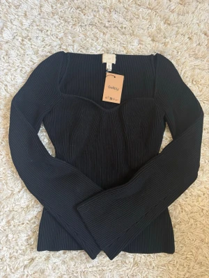 Svart ribbad topp från H&M - Snygg svart ribbad topp från H&M med markerad byst och lång ärm. Toppen har en figurnära passform och fyrkantig ringning som ger en modern look. Perfekt att styla med jeans eller kjol för en trendig outfit.