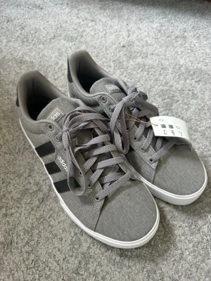 Grå Adidas Daily 3.0 - Snygga grå sneakers från Adidas med klassiska svarta ränder på sidan och vit sula. Skorna har textilovandel, matchande grå skosnören och låg profil. Perfekta för en clean och sportig look.