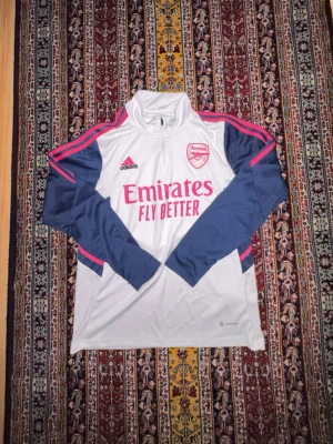Arsenal träningströja Adidas vit/blå - Snygg långärmad Arsenal träningströja från Adidas i vitt med marinblå ärmar och rosa detaljer. Tröjan har halv dragkedja, klubbmärke och Adidas-logga på bröstet samt 'Emirates Fly Better' tryckt framtill. Tillverkad i funktionsmaterial som andas.