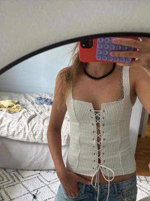 Vit randig korsettopp med snörning - Säljer en vit korsettopp med tunna svarta ränder och snygg snörning framtill. Toppen har breda axelband och en croppad passform som ger en edgy vibe. Perfekt att styla med jeans eller kjol för en trendig look.