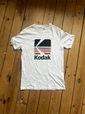 Vit Kodak t-shirt med tryck - Säljer en vit t-shirt med stort retro Kodak-tryck på framsidan och texten 'A Kodak Moment.' på ryggen. Klassisk passform och rund hals. Perfekt för dig som gillar vintage vibes och grafiska prints. Har blivit lite gul under ene ärmen och på innersidan av halsringen baktil, men inget som är särskilt stort.