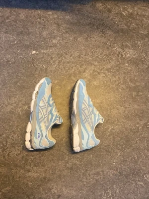 Blåvita Asics sneakers - Säljer ett par fräscha sneakers från Asics i ljusblått och vitt. Skorna har meshpaneler för bra ventilation, detaljer i mocka och syntet samt klassisk snörning. Sulan är vit med grå inslag och ger bra stöd. Perfekta för en sportig och trendig look.