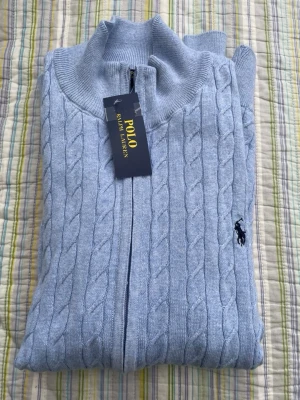Ljusblå kabelstickad ziptröja Polo - Ljusblå kabelstickad ziptröja från Polo Ralph Lauren med hög krage och dragkedja framtill. Tröjan har det klassiska broderade logotypen på bröstet och ribbade muddar. Perfekt för en stilren och avslappnad look.