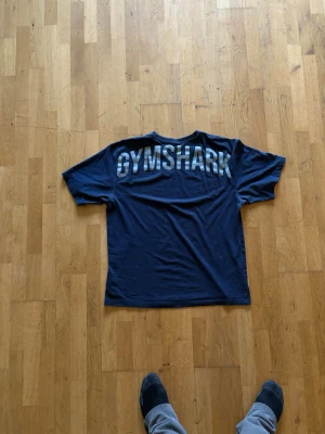Blå Gymshark t-shirt med tryck - Mörkblå t-shirt från Gymshark med stort, grått tryck över ryggen. Klassisk rund hals och korta ärmar. Tillverkad i mjuk bomull som känns skön mot huden. Perfekt för dig som gillar sportig och enkel stil.