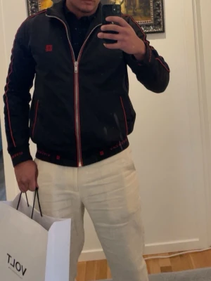 Givenchy Svart tröja med dragkedja - Snygg svart ziptröja från Givenchy med röda detaljer längs dragkedja, ärmar och fickor. Tröjan har hög krage och ribbade muddar. Perfekt för dig som gillar sportig och stilren look.
