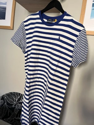 Randig t-shirt klänning från Polo Ralph Lauren - Snygg randig t-shirt klänning från Polo Ralph Lauren i blått och vitt. Klassisk passform med rund hals och korta ärmar. Tillverkad i mjuk bomull och har en liten broderad logga på bröstet. Perfekt för en avslappnad och stilren look.