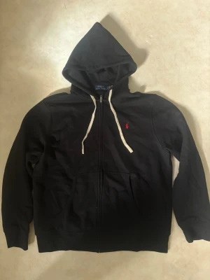 Ny oanvänd Svart hoodie från Polo Ralph Lauren - Svart hoodie från Polo Ralph Lauren med dragkedja, vit dragsko och klassisk röd logga på bröstet. Tillverkad i mjukt bomullsmaterial och har två fickor framtill. Perfekt för en avslappnad och stilren look. Ny och oanvänd