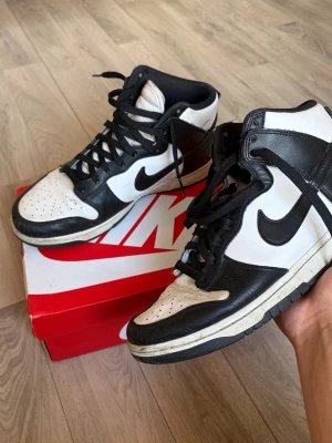 Nike Dunk High svart/vit sneakers - Nike Dunk High sneakers i svart och vitt läder med klassisk Swoosh på sidan. Skorna har högt skaft, svart snörning och vit tåbox med perforeringar. Tidlös och clean design som passar till det mesta. Kommer med originalkartong.
