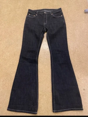 Svarta jeans med vita sömmar  - Säljer ett par svarta bootcut jeans från Weekday med klassisk femficksmodell och kontrastsömmar. Jeansen har low waist och vida ben, perfekta för en trendig och avslappnad look. Tillverkade i kraftig denim med snygg passform. Aldrig använda, passar även strl M
