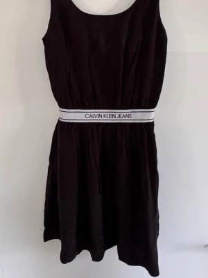Svart klänning Calvin Klein Jeans - Svart kortklänning från Calvin Klein Jeans med bred, vit resår i midjan där loggan står tryckt. Klänningen är ärmlös med rund halsringning och har en enkel, sportig look. Perfekt för dig som gillar stilrena och bekväma plagg.