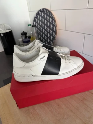 Vita sneakers med svart detalj - Snygga vita sneakers med en bred svart remsa över mitten. Skorna har låga skaft, vita snören och en klassisk siluett. Tillverkade i slätt läder för en clean look som passar till det mesta. Perfekta för dig som gillar stilrena och moderna sneakers. Köpta på ILLUM i Danmark för 4750 danska som är ca 7000 svenska kr. Mitt pris 3500 kr. Finns i Malmö så kan hämtas men kan också skickas.