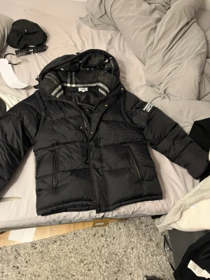 Svart pufferjacka M - Säljer en svart pufferjacka med stor huva och grått, rutigt innerfoder. Jackan har dragkedja, knappar och två snedställda fickor framtill. Tydlig patch på ärmen och klassisk design som håller dig varm under kalla dagar.