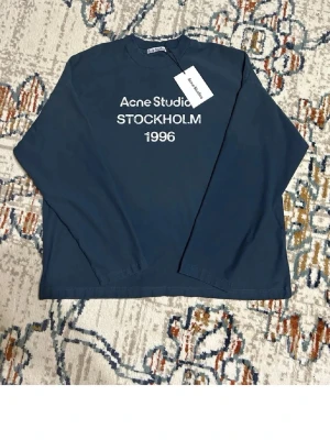 Blå långärmad topp Acne Studios - Mörkblå långärmad topp från Acne Studios med vit text 'Acne Studios STOCKHOLM 1996' på bröstet. Klassisk rund halsringning och avslappnad passform. Perfekt för en stilren och trendig look. Tvättlappen är avklippt, och även lapparna på bilden är avklippta, kvitto finns