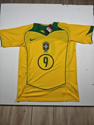 Brasilien Ronaldo 9 matchtröja Nike - Gul Brasilien fotbollströja från Nike med gröna detaljer, nummer 9 och Ronaldo på ryggen. Tröjan har korta ärmar, rund hals och Brasilien-emblem på bröstet. Perfekt för dig som älskar fotboll och vill hylla en ikonisk spelare.