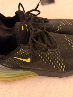 Nike Air Max 270 svarta sneakers - Nike Air Max 270 sneakers i svart med gula detaljer och synlig Air-sula. Skorna har snörning och meshmaterial för bra ventilation. Perfekta för dig som vill ha en sportig och modern look.i absolut nyskick