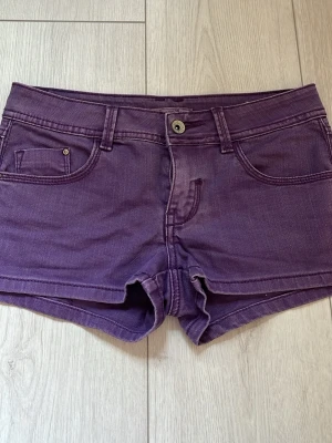 Lila denimshorts med fickor - Säljer ett par lila denimshorts med klassisk femficksdesign och knappgylf. Shortsen har bälteshällor och snygga bakfickor med dekorativa sömmar. Perfekta för varma dagar och ger en cool vibe till din outfit.