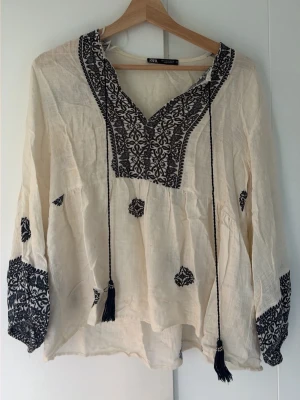 Bohemisk blus med brodyr från Zara - Krämvit boho-blus från Zara med svarta broderier och dekorativa tofsar. Långärmad modell i tunn, luftig bomull med v-ringning och breda broderade muddar. Perfekt för en avslappnad och trendig look. Den har ett jättelitet hål på baksidan.