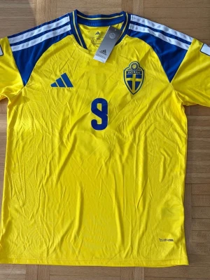 Sverige fotbollströja gul Adidas - Gul fotbollströja från Adidas med blå detaljer och nummer 9 på bröstet. Tröjan har Sveriges landslagsemblem på vänster sida och Adidas-logga på höger. Tillverkad i lätt och ventilerande material med ClimaCool-teknologi. Perfekt för match eller träning.