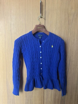 Blå kabelstickad kofta med volang - Supersnygg blå kabelstickad kofta från Ralph Lauren med volangkant nedtill och vita knappar framtill. Klassisk gul logga broderad på bröstet. Långärmad och rundhalsad modell som ger en preppy känsla.