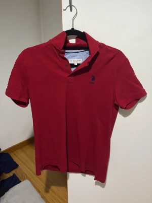 Röd pikétröja från U.S. Polo Assn. - Snygg röd pikétröja från U.S. Polo Assn. med klassisk krage och korta ärmar. Tröjan har en liten mörkblå logga broderad på bröstet och är tillverkad i mjuk bomull. Perfekt till jeans eller shorts för en avslappnad look.