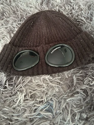 Cp company mössa svart  - Säljer en brun ribbstickad mössa från C.P. Company med ikoniska goggles framtill. Mössan har en uppvikt kant och är tillverkad i ett mjukt, stretchigt material som sitter skönt på huvudet. Perfekt för dig som vill ha en streetwear-inspirerad look.