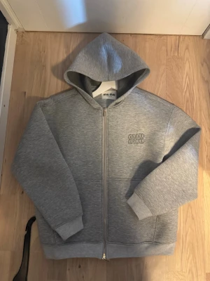 Grå hoodie med dragkedja från Miu Miu - Säljer en stilren grå hoodie från Miu Miu med dragkedja framtill och broderad logga på bröstet. Hoodien har huva, ribbade muddar och två fickor framtill. Tillverkad i mjukt sweatshirtmaterial som är perfekt för en avslappnad look.