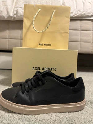 Svarta Axel Arigato sneakers - Stilrena svarta sneakers från Axel Arigato med lågt skaft och klassisk siluett. Skorna har svart ovandel i läder, svarta skosnören och vit sula. Perfekta för dig som gillar minimalistisk och clean design.