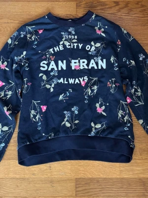 Blommig sweatshirt från H&M - Mörkblå sweatshirt från H&M med trycket ”The City of San Fran Always” framtill. Tröjan har ett blommigt mönster i rosa och blått samt ribbade muddar vid ärmslut och nederkant.